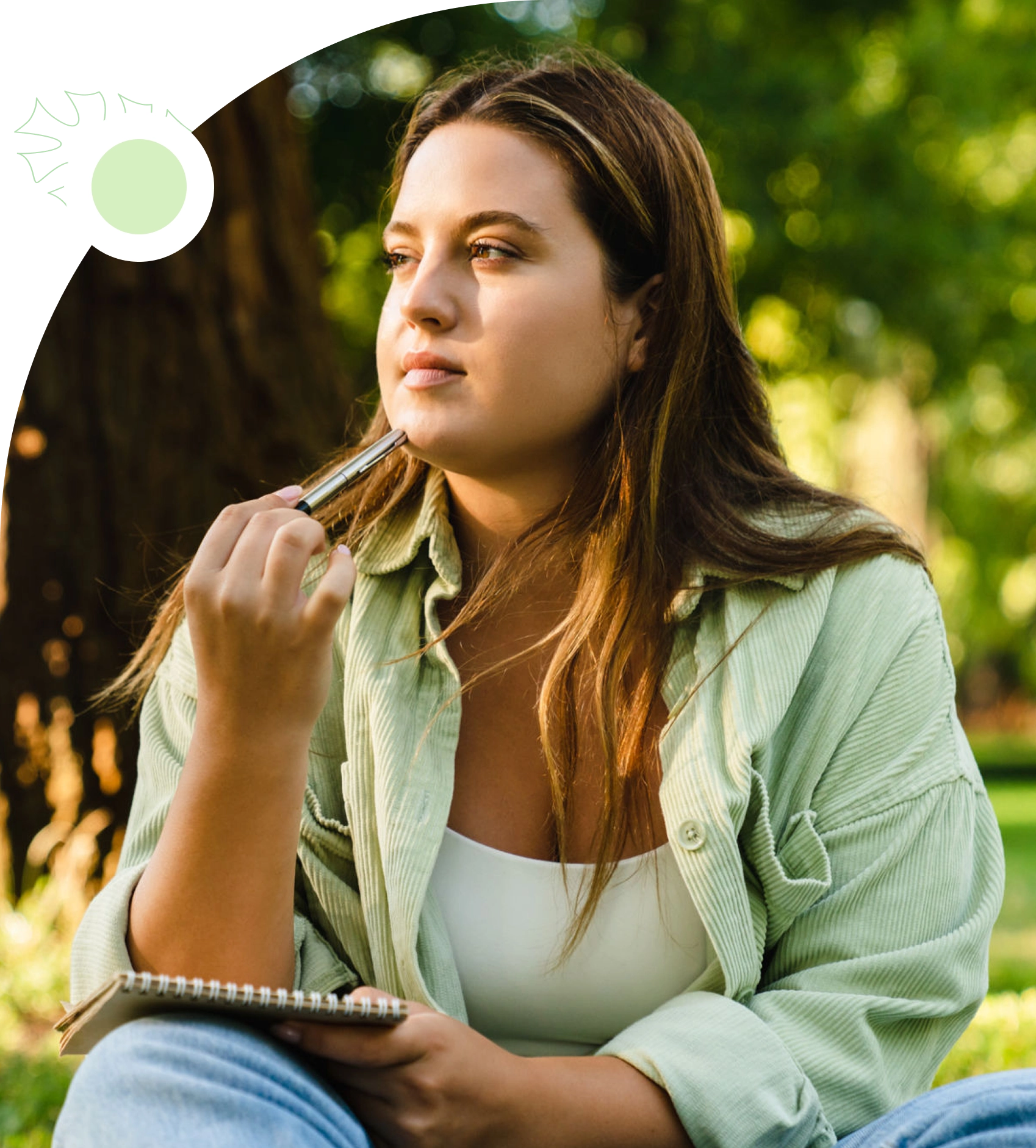 A person sits outdoors on grass, holding a pen to their chin and looking thoughtful, with a notebook in hand. Trees and greenery are visible in the background.